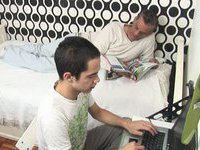 Old guy lies on the bed seductively and reads magazine while his young lover enjoys playing his computer